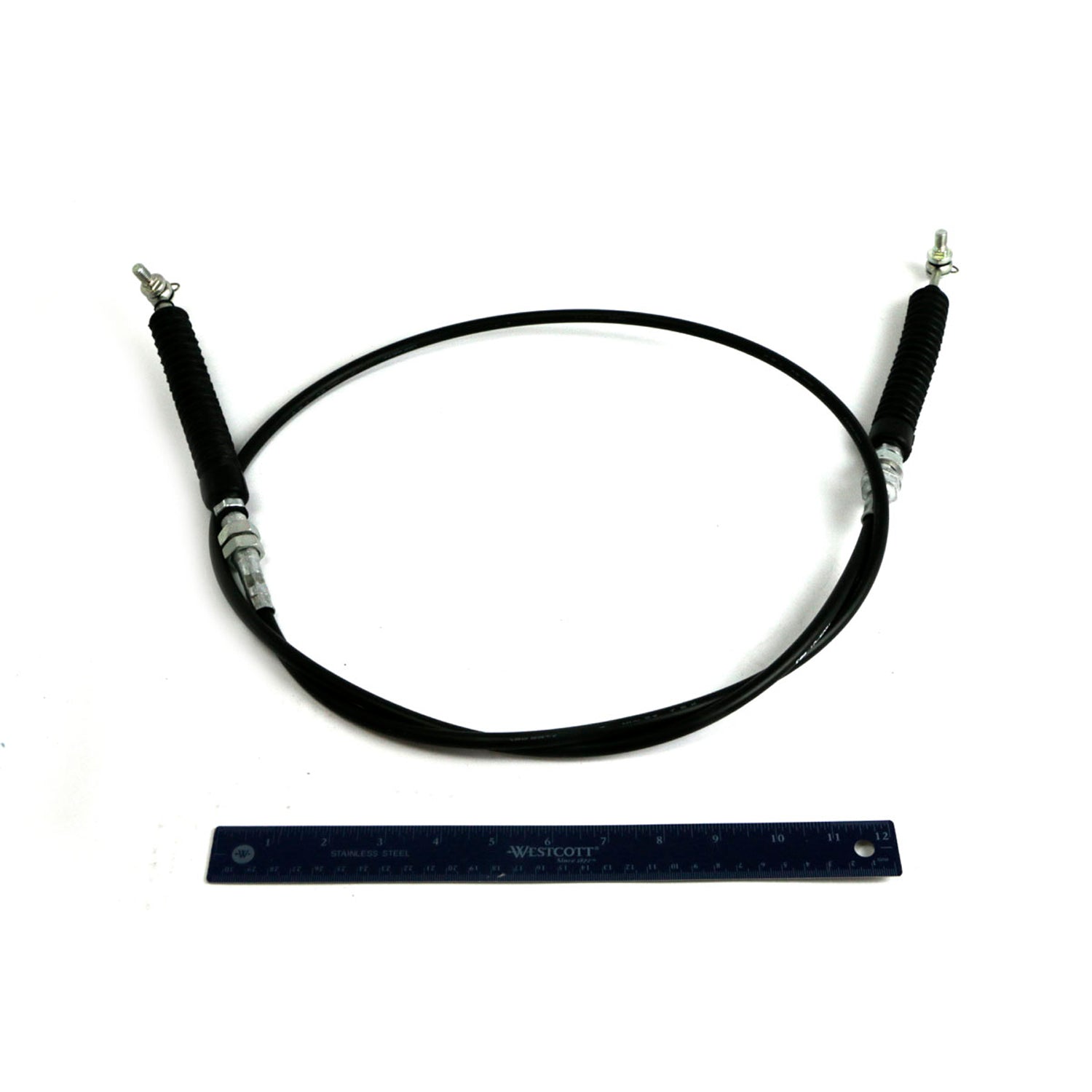 Black flexible shift cable with metal fittings on both ends and protective sleeves near connectors, shown with a blue ruler.