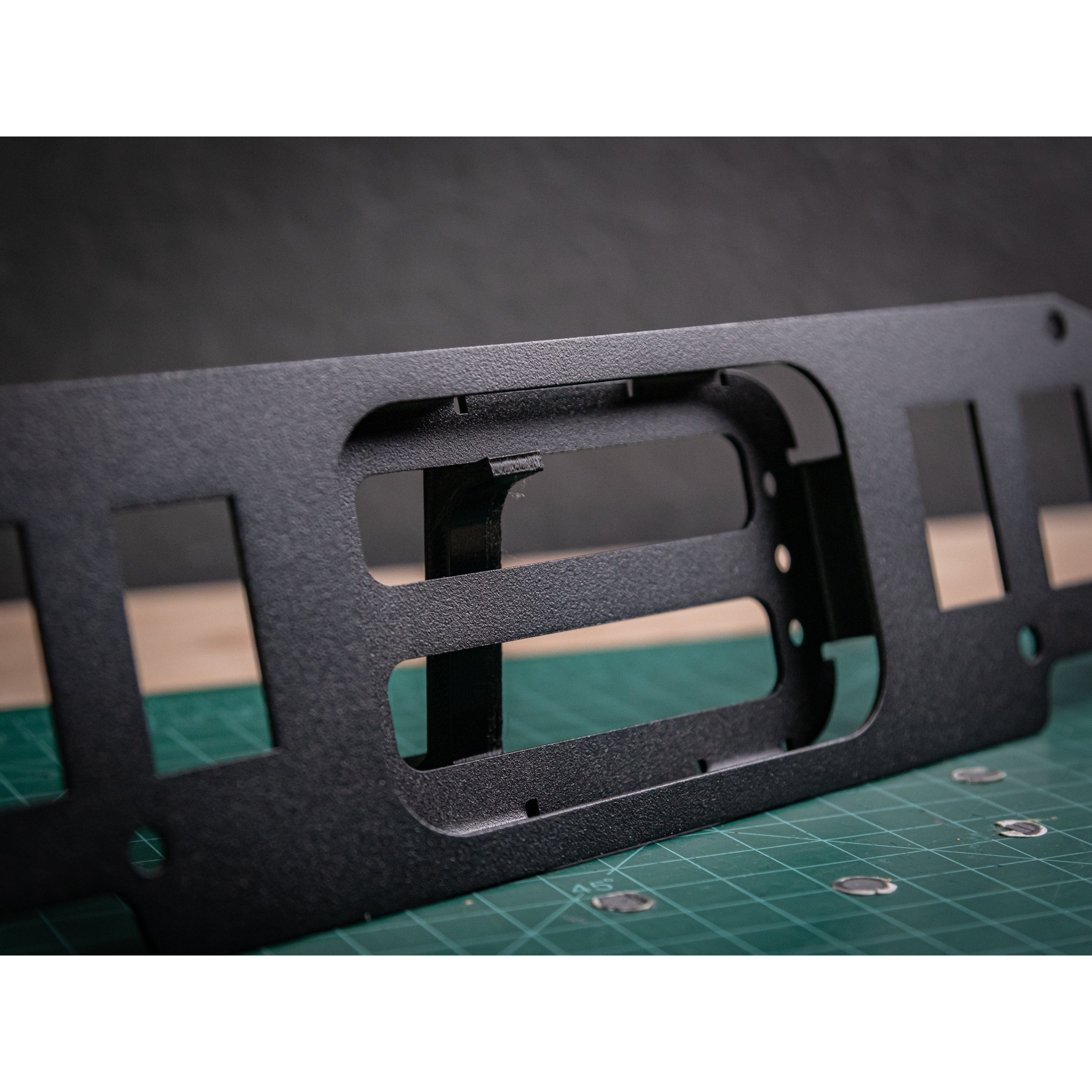 Close-up of the Turbo S Auxbeam 8 Gang Mount by StayFlush, a black metal bracket with rectangular cutouts, ideal for Polaris Turbo S or Auxbeam Switch Panel. Shown on a green cutting mat with measurement lines and screws, blurred dark background.