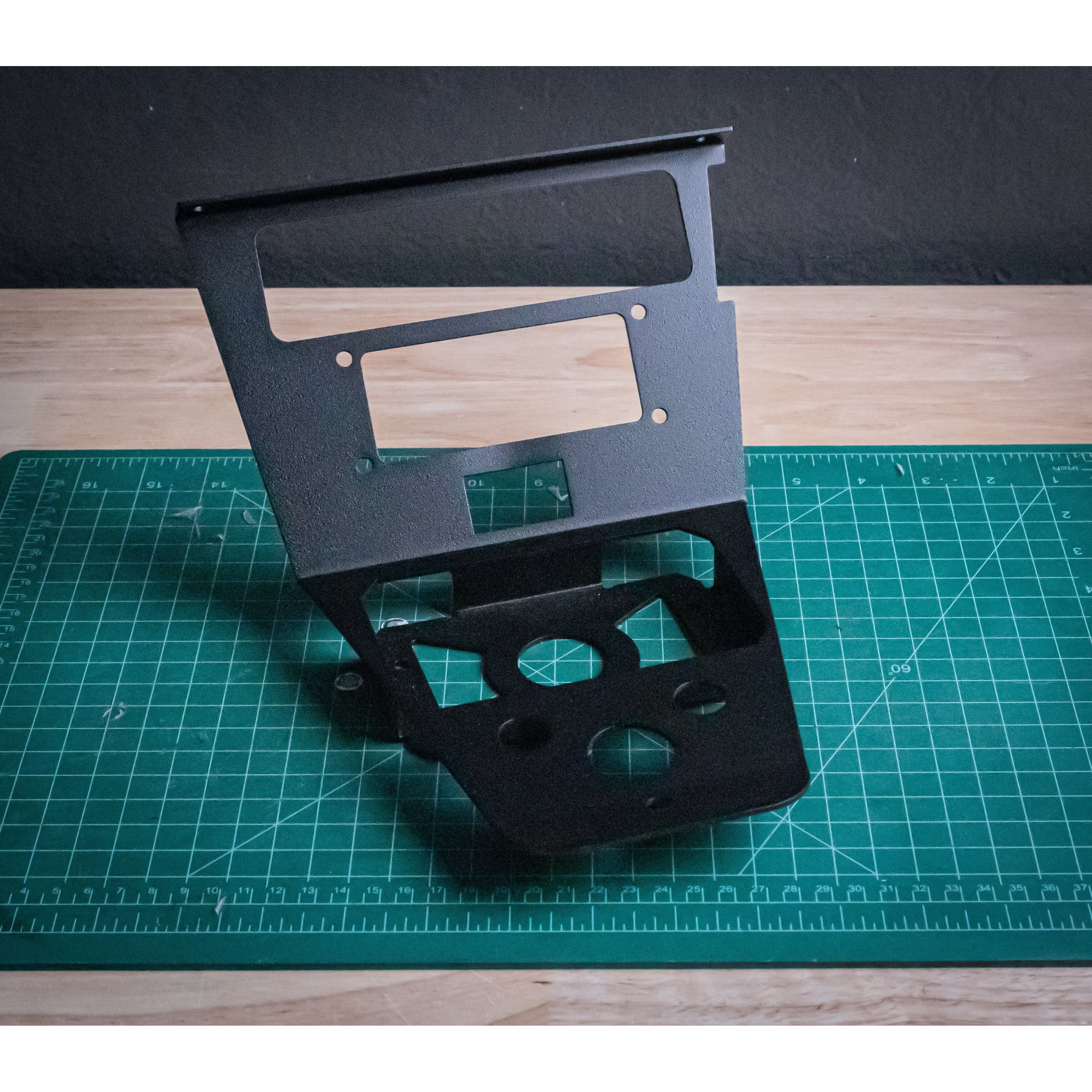 A black PCI Kenwood Switchworks Mount by StayFlush with various cutouts and angles stands upright on a green cutting mat atop a wooden table, designed for seamless radio integration in Polaris Pro/ProR vehicles.
