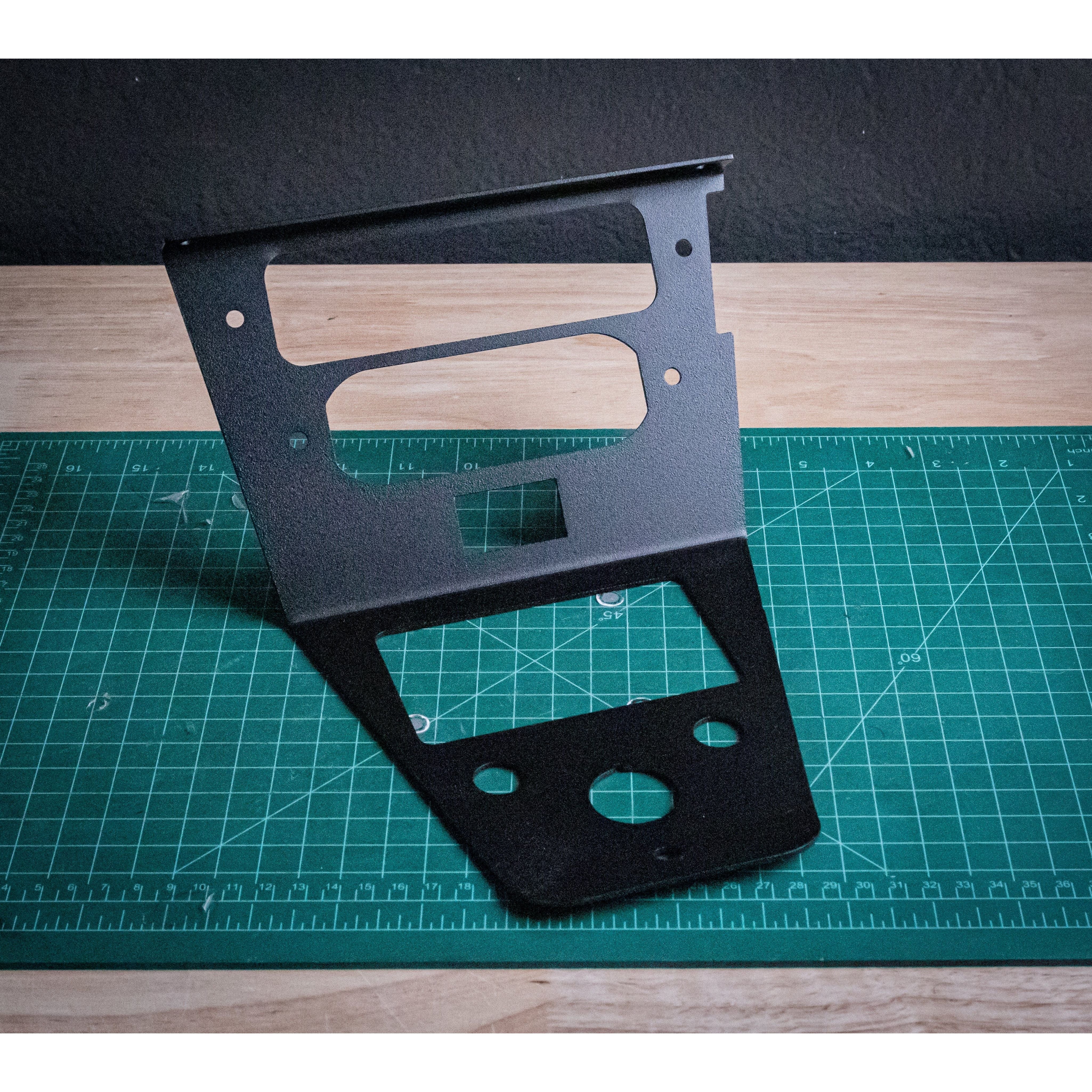 The Rugged Radio RLB Mount by StayFlush is a black metal dashboard panel with cutouts for a Rugged Radio mount and rocker switch, shown upright on a green cutting mat with grid lines on a wooden surface.