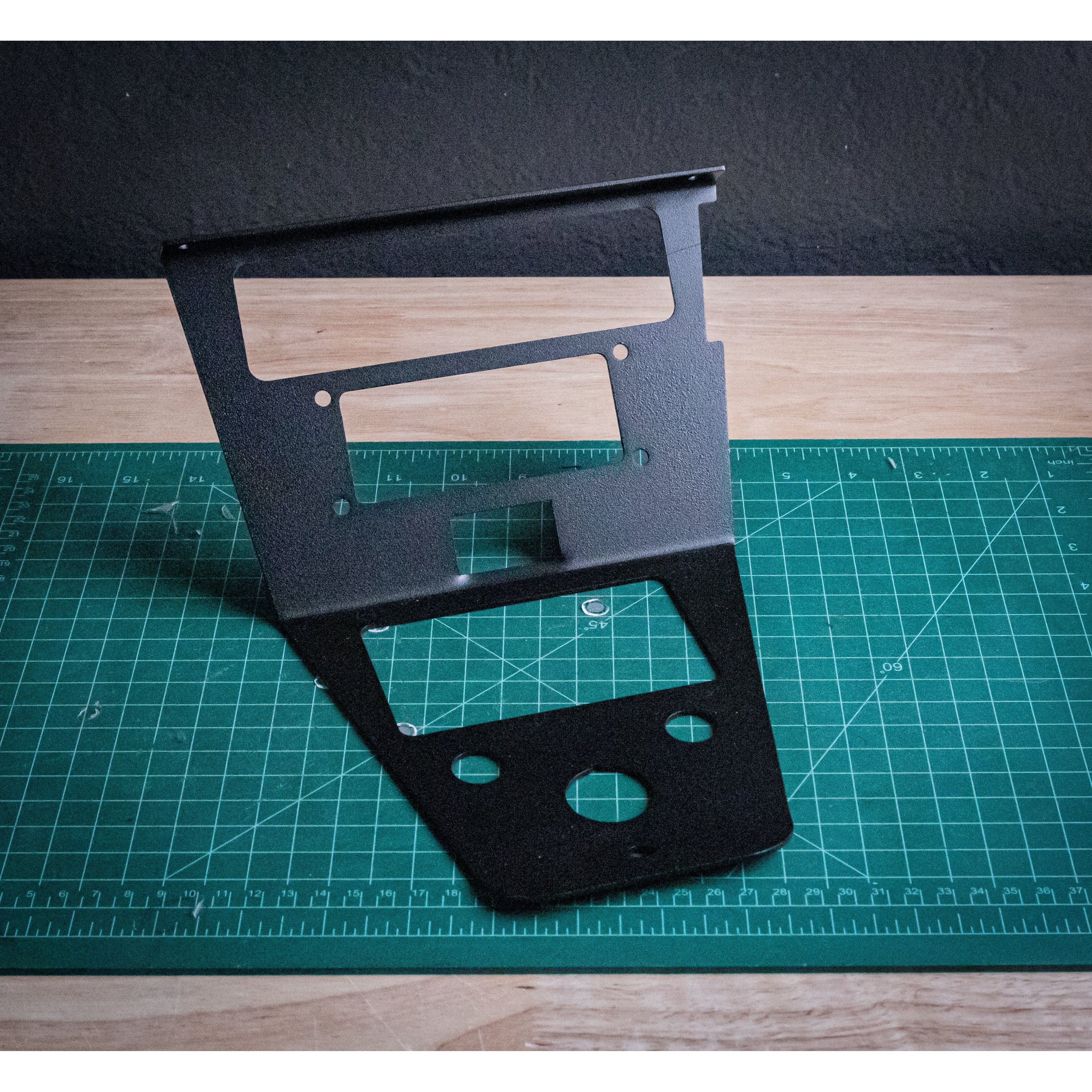 The PCI Kenwood RLB Mount by StayFlush, a black metal dashboard panel with multiple cutouts and Polaris Pro Compatibility, stands upright on a green cutting mat atop a wooden surface against a dark wall.