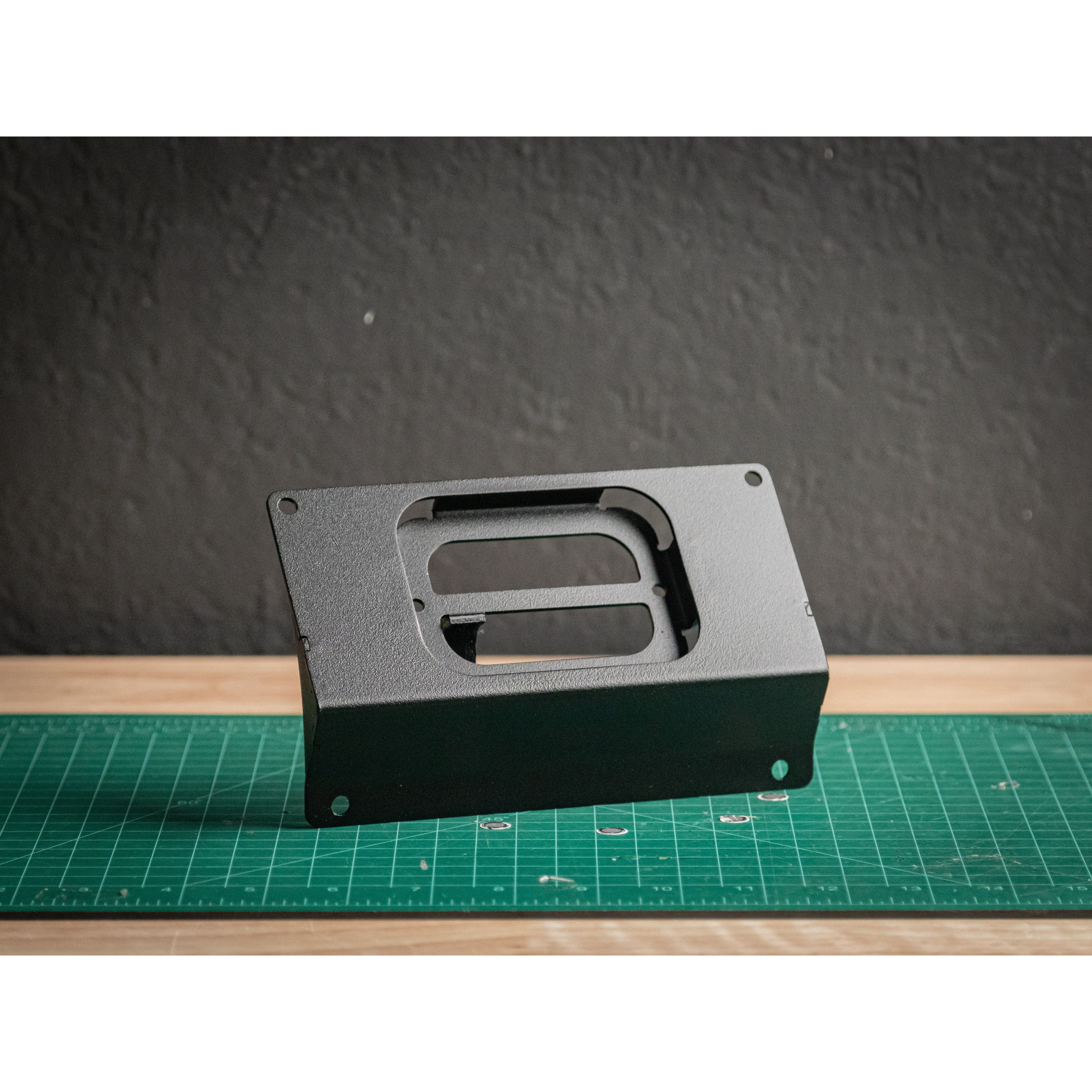 The Polaris 200 EFI Auxbeam 6 Mount by StayFlush, a black rectangular metal bracket, stands upright on a green grid cutting mat. Behind it is a textured dark wall and a wooden surface.