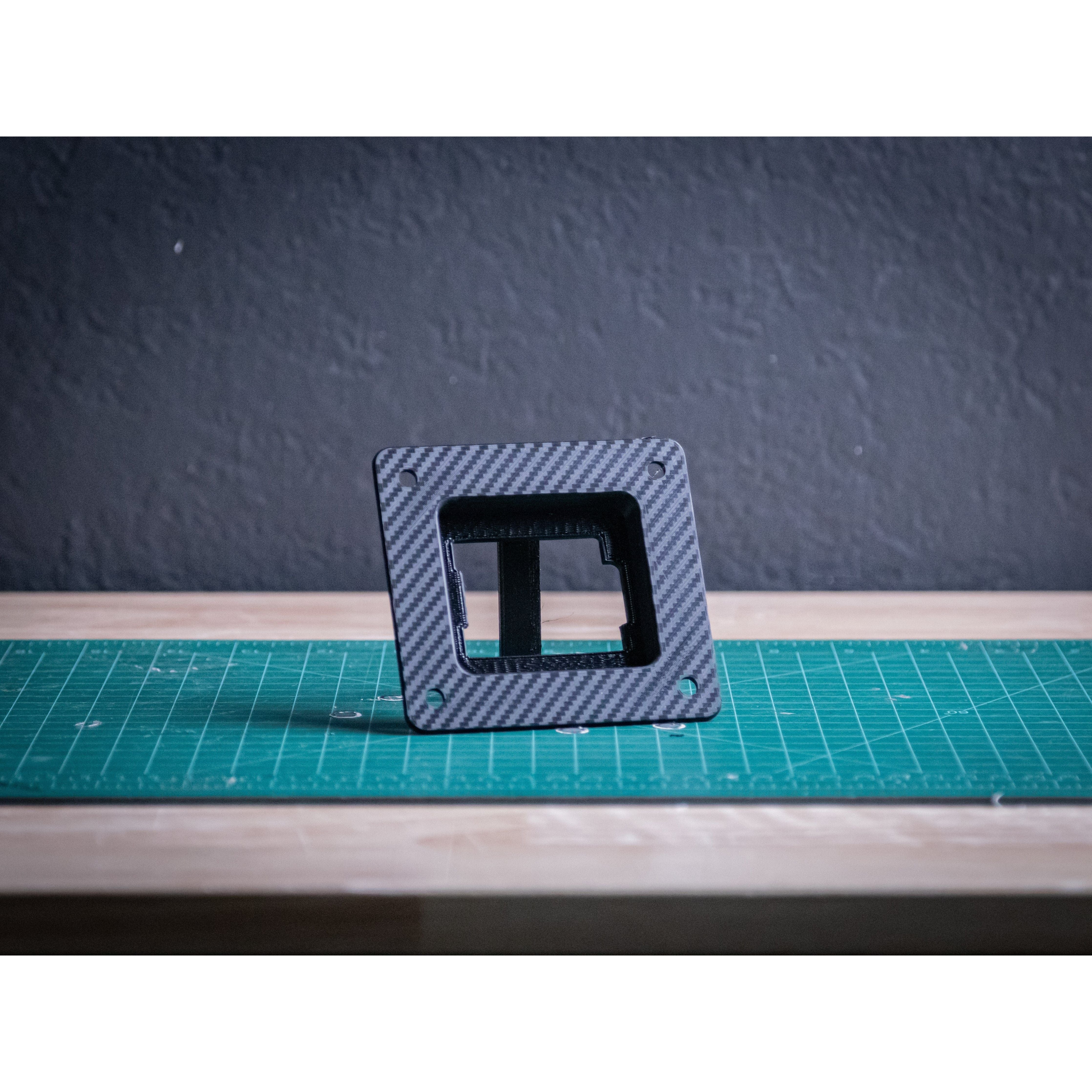 The Can-am X3 DPS Bezel by StayFlush, featuring a custom dash design and hollow center, stands upright on a green cutting mat against a dark textured wall, highlighting its premium carbon fiber finish.