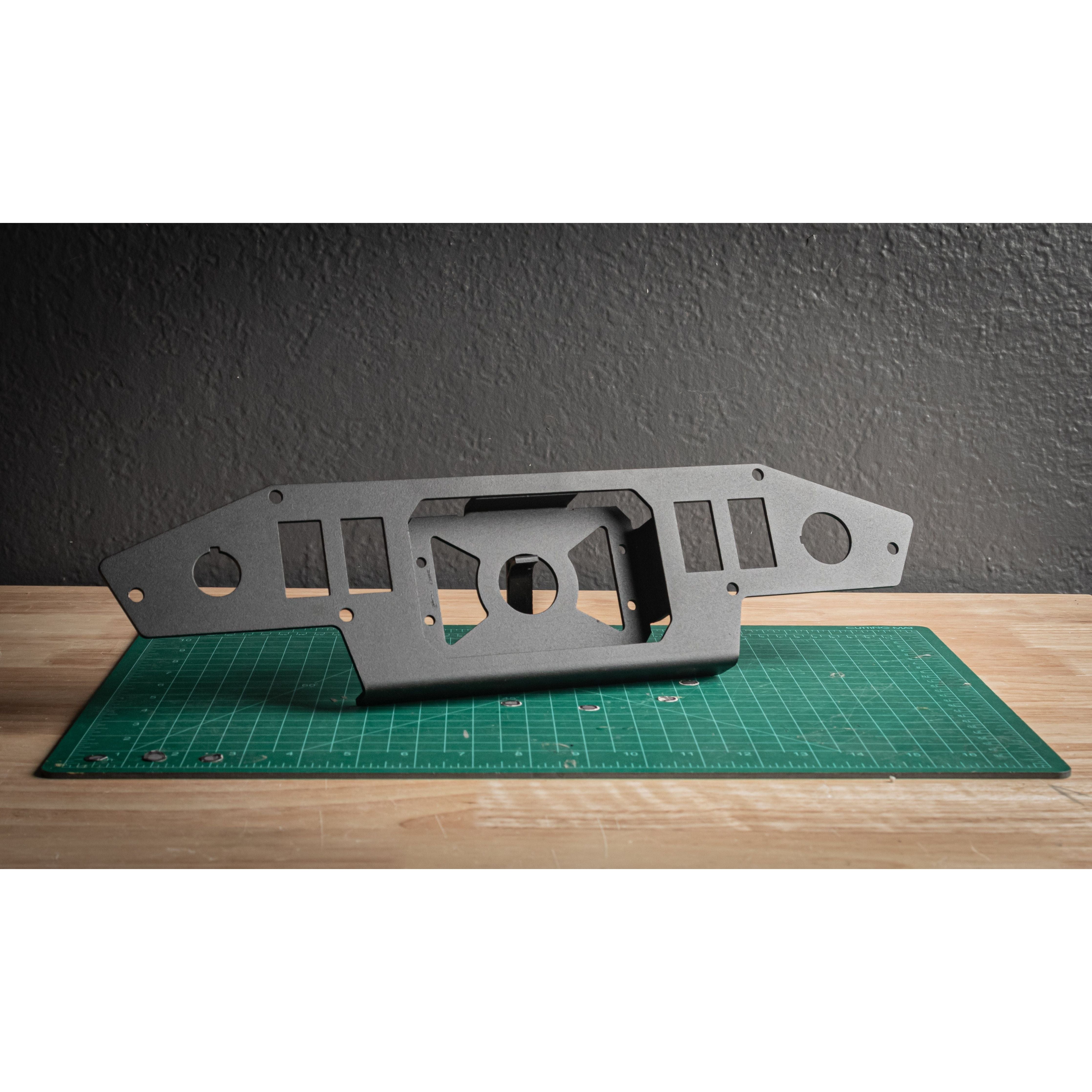 A flat, black Turbo S Alpha Switchworks Mount by StayFlush with multiple cutouts and rocker switch slots stands upright on a green cutting mat on a wooden table, against a textured gray wall.