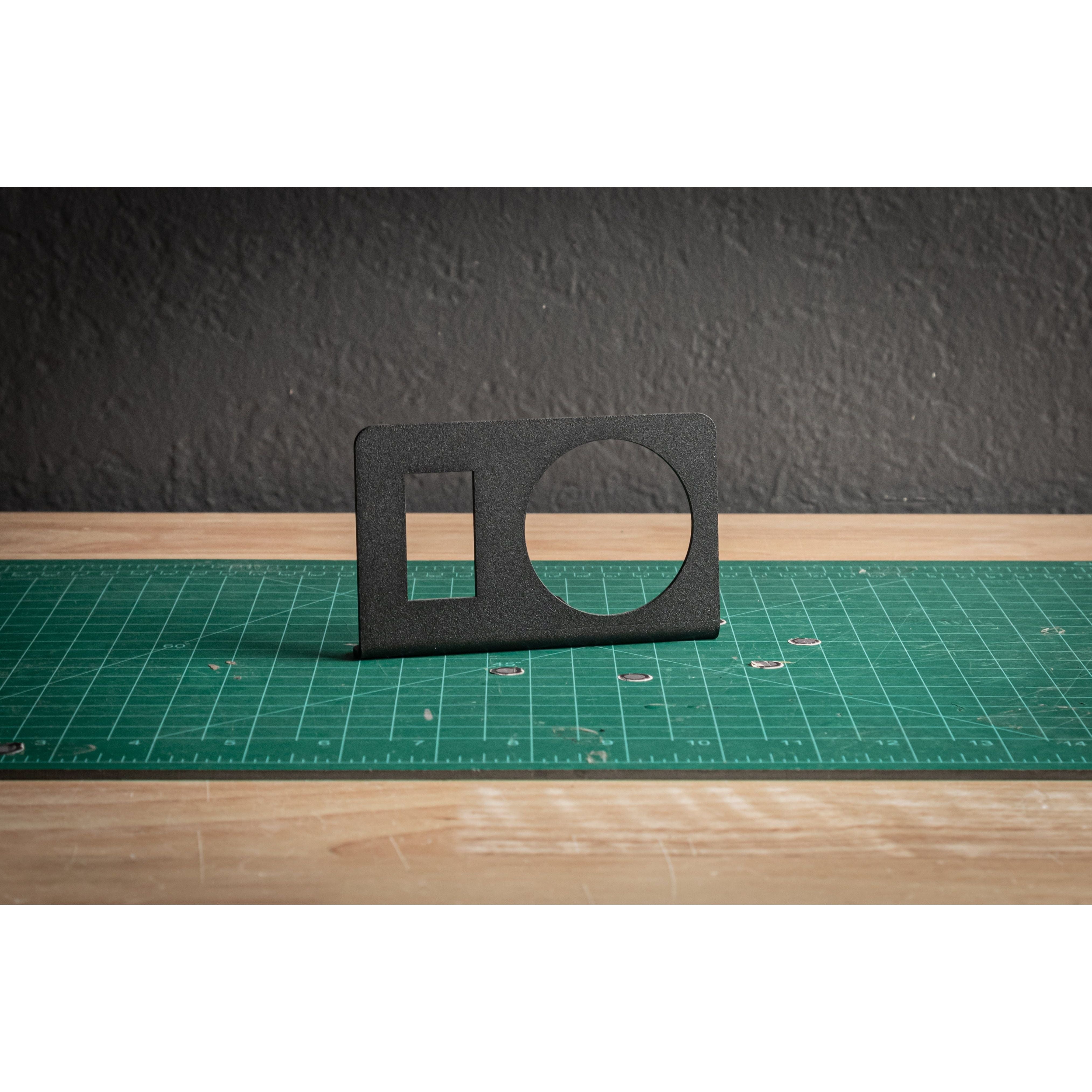 The Rocker+Gauge Sidekick by StayFlush is a black rectangular mount with a large circular and small square cutout, shown upright on a green mat—ideal for flat surface mount & CanamX3 use, with textured gray wall and wood background.