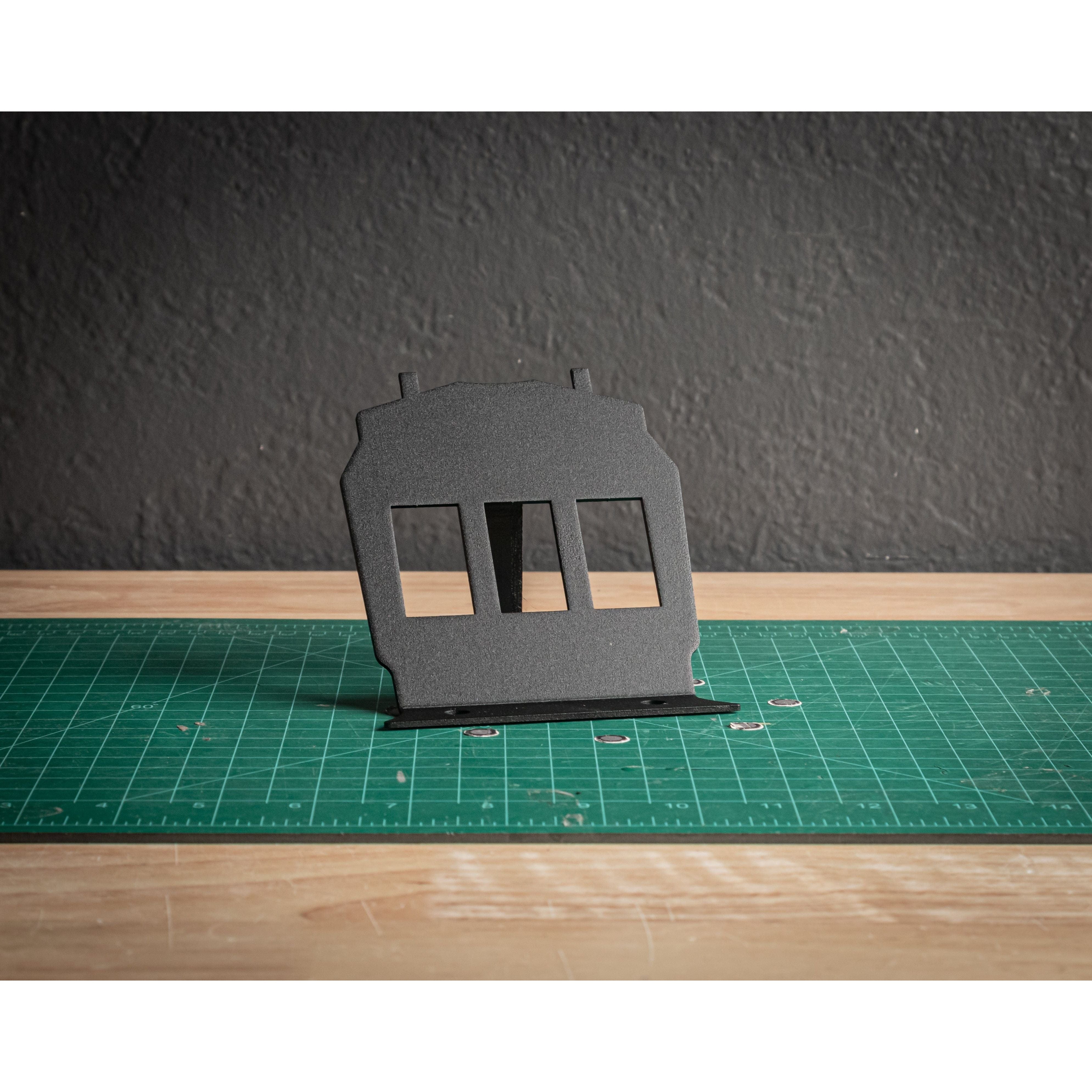 A StayFlush Rocker Switch Lower Plate by StayFlush, a black window-shaped aluminum mount, stands upright on a green cutting mat on a wooden table with a dark textured wall in the background.