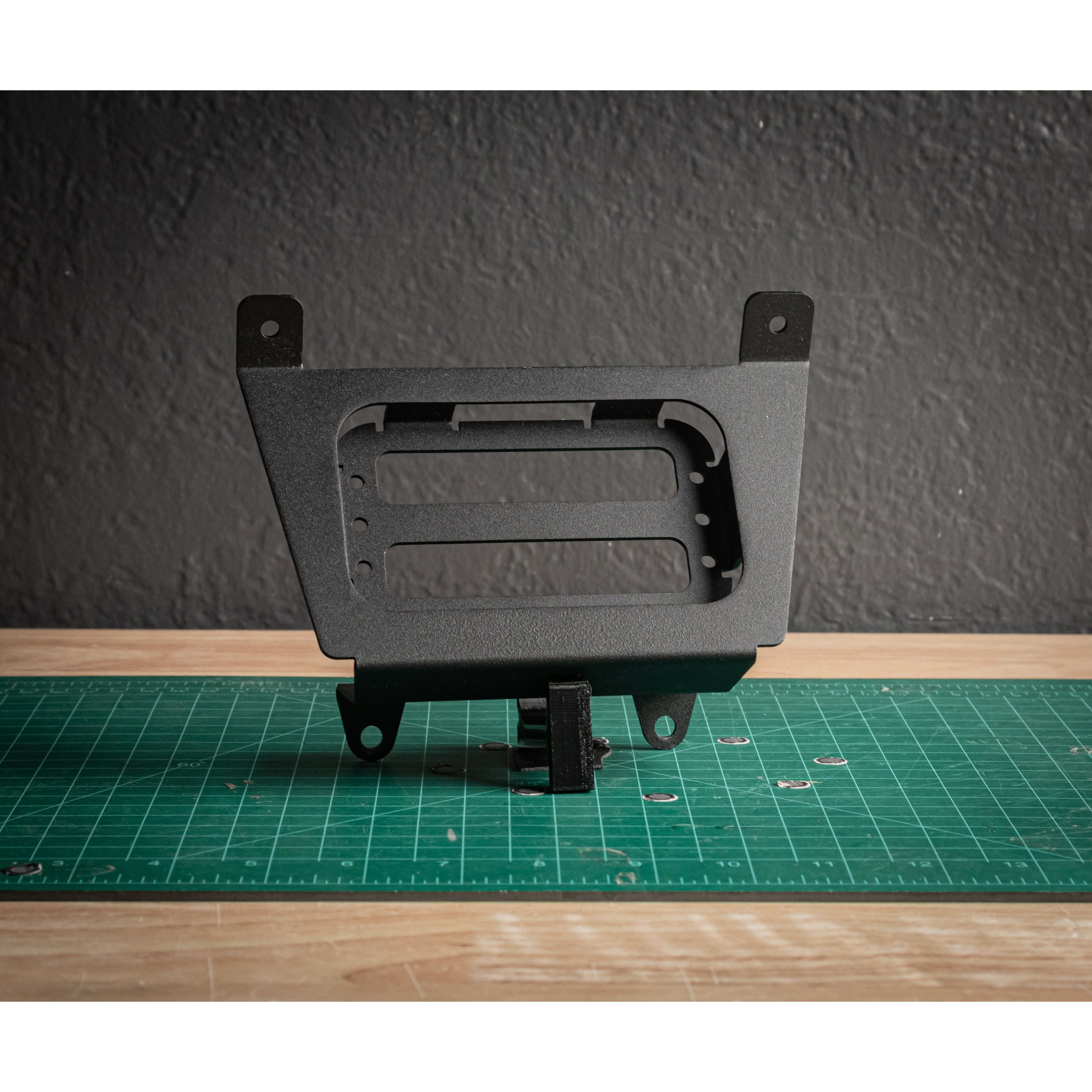 A black metal mount bracket for the Pro/R Auxbeam 6 Gang by StayFlush stands upright on a green cutting mat atop wood, with a dark gray wall behind—perfect for 12v Auxbeam installations.