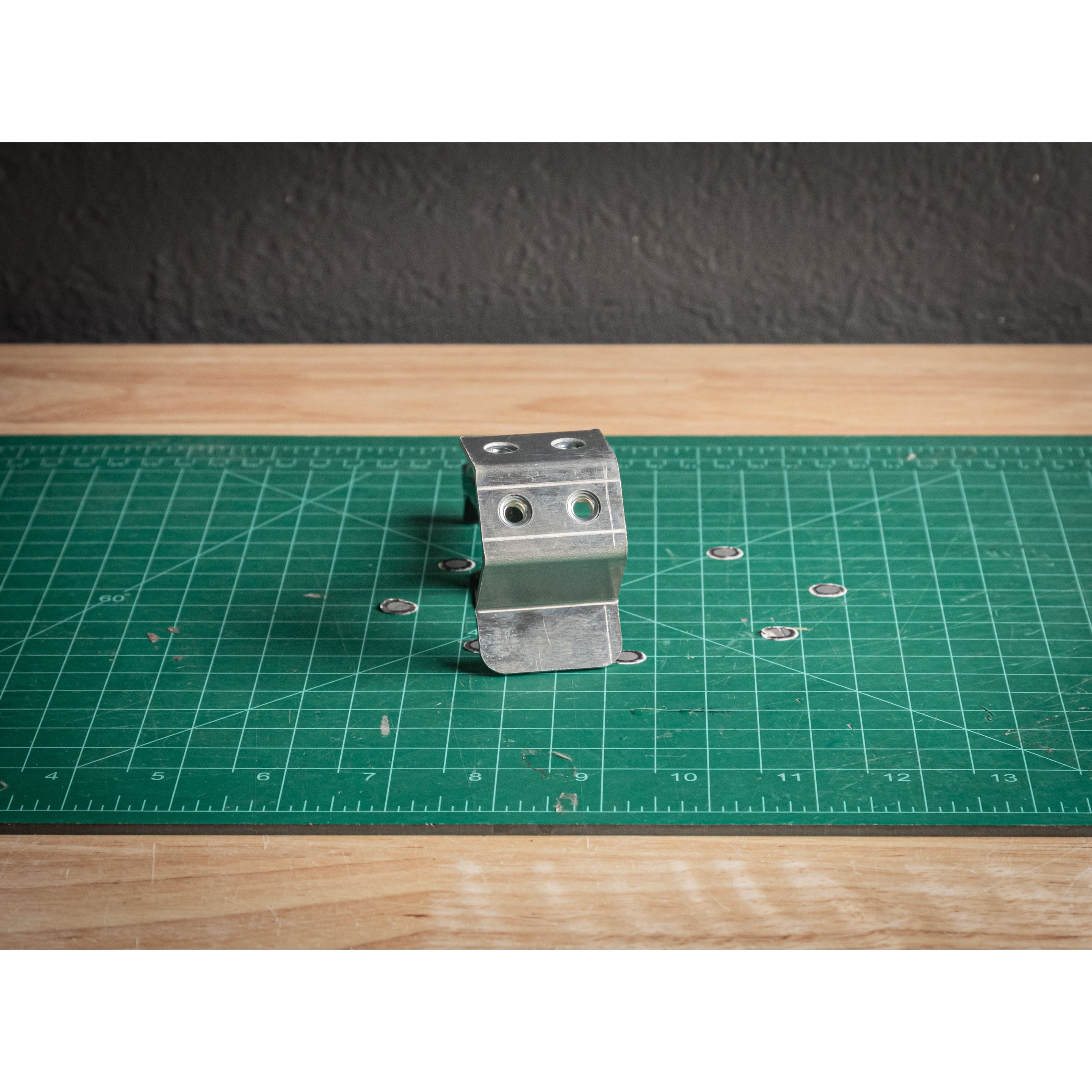 The Terminal Bus Post by StayFlush, a metal bracket with four holes perfect for battery terminal mounts, stands upright on a green cutting mat with metal washers nearby. The scene features a wooden surface and dark textured wall in the background.