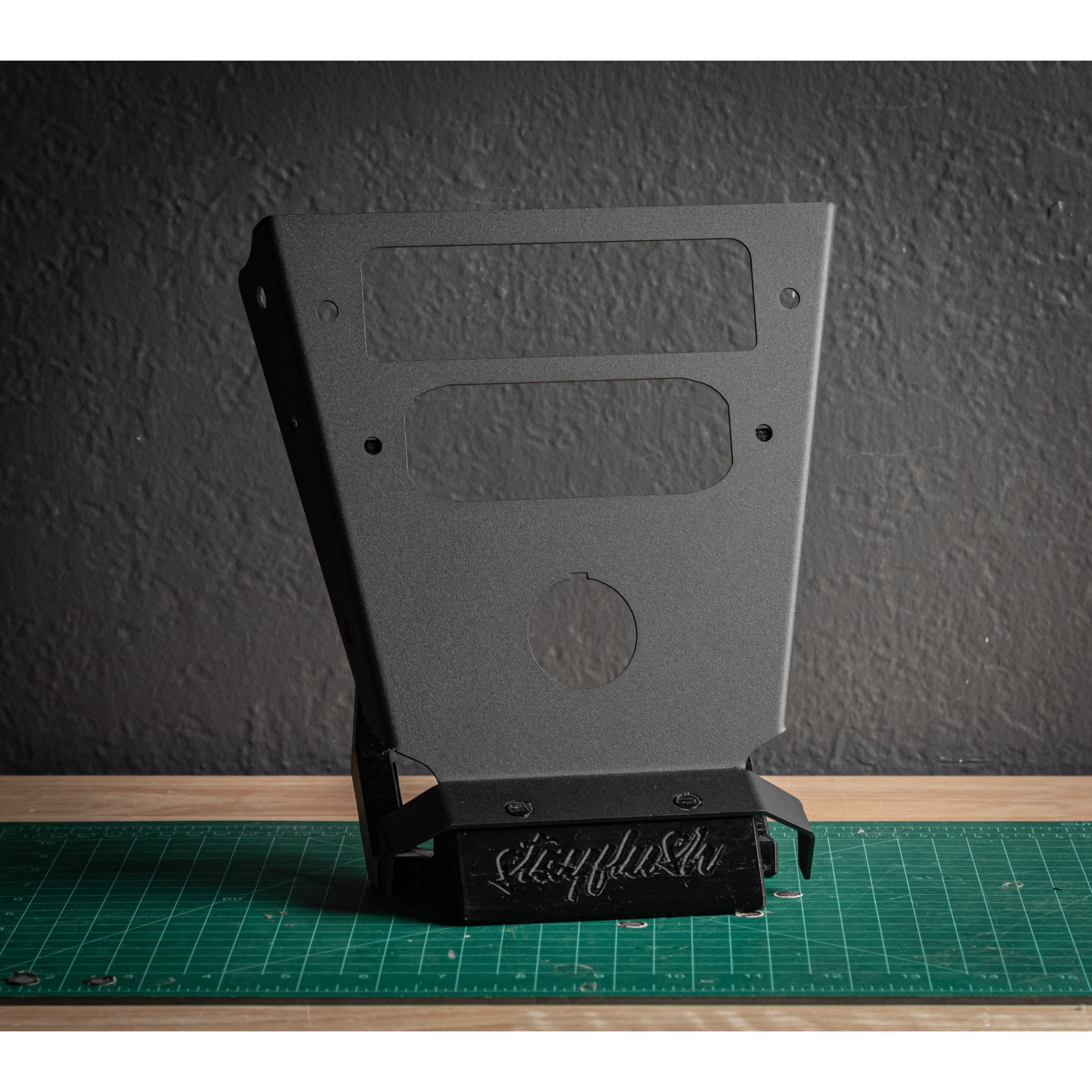 The Rugged Radio Mount/RM60 (Classic) by StayFlush is a black metal bracket featuring cutouts and stayflush engraved at the base, shown standing on a green cutting mat against a textured dark gray wall.