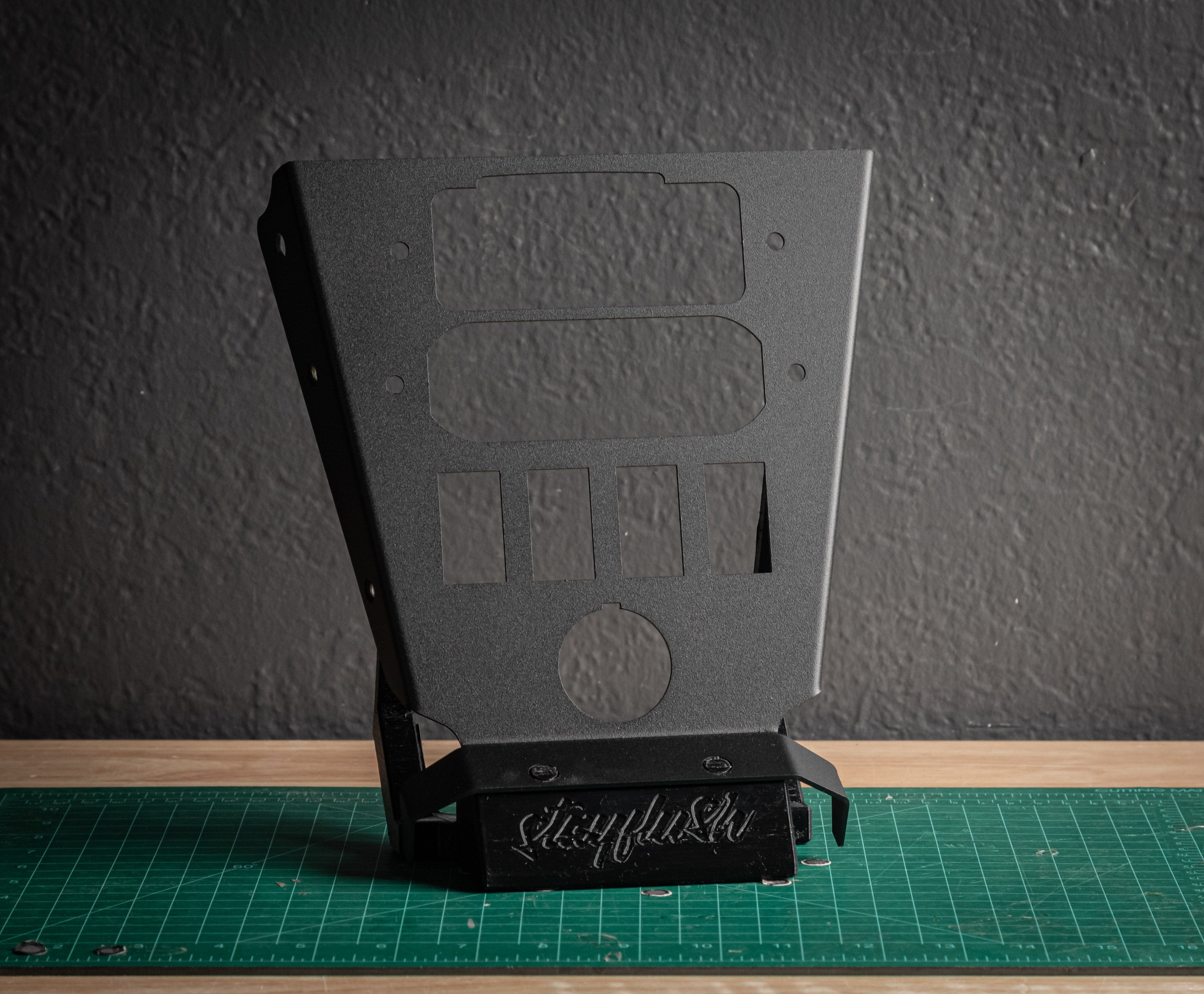 The ABM Mini Mount Plus 4 by StayFlush is a matte black metal bracket with multiple cutouts, standing vertically on a green cutting mat before a textured gray wall. sloppyfabs is engraved at its flush-mount base.
