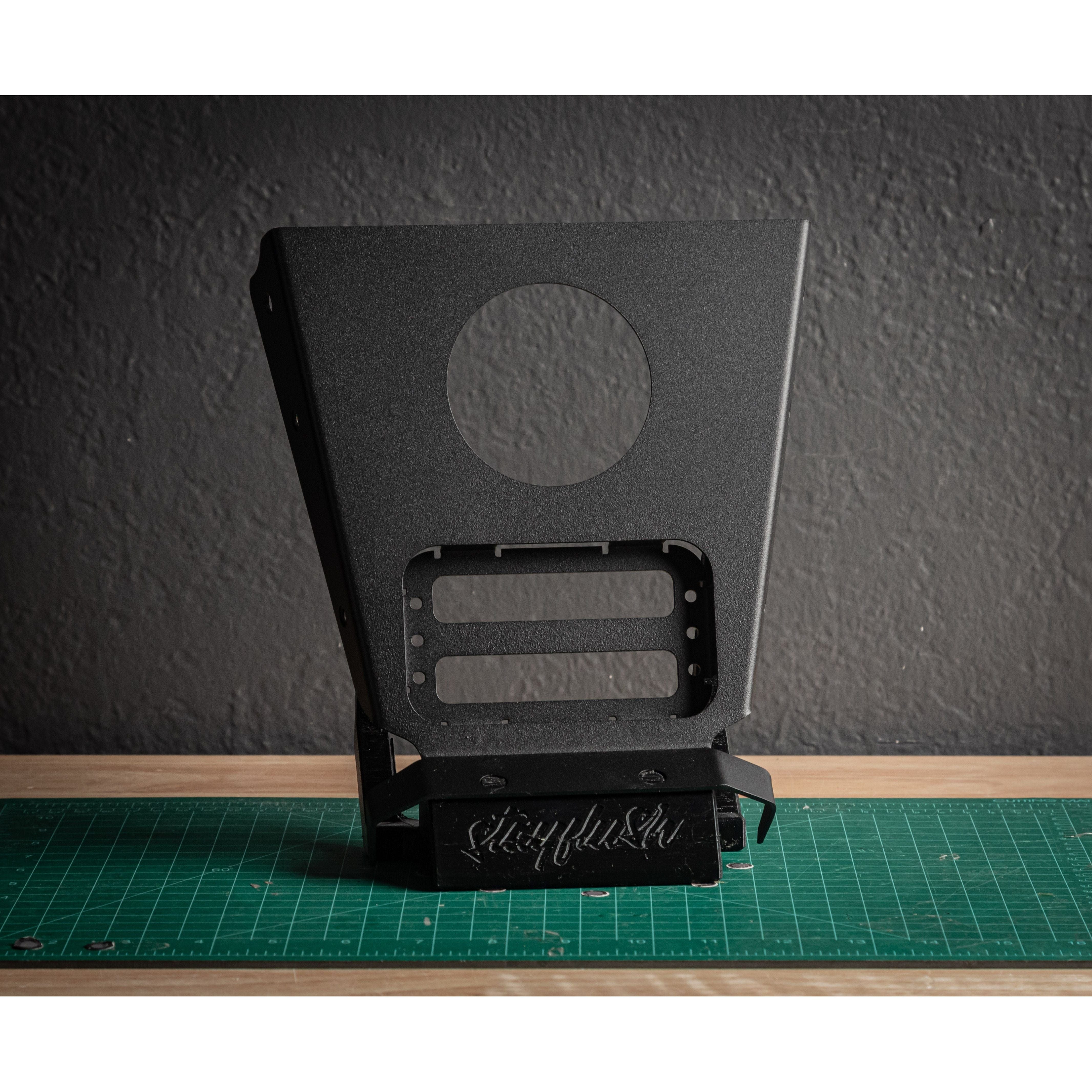A StayFlush Cyclops/Auxbeam Mount by StayFlush, a black metal bracket with a large circular hole and rectangular cutouts, stands upright on a green cutting mat against a textured dark gray wall.