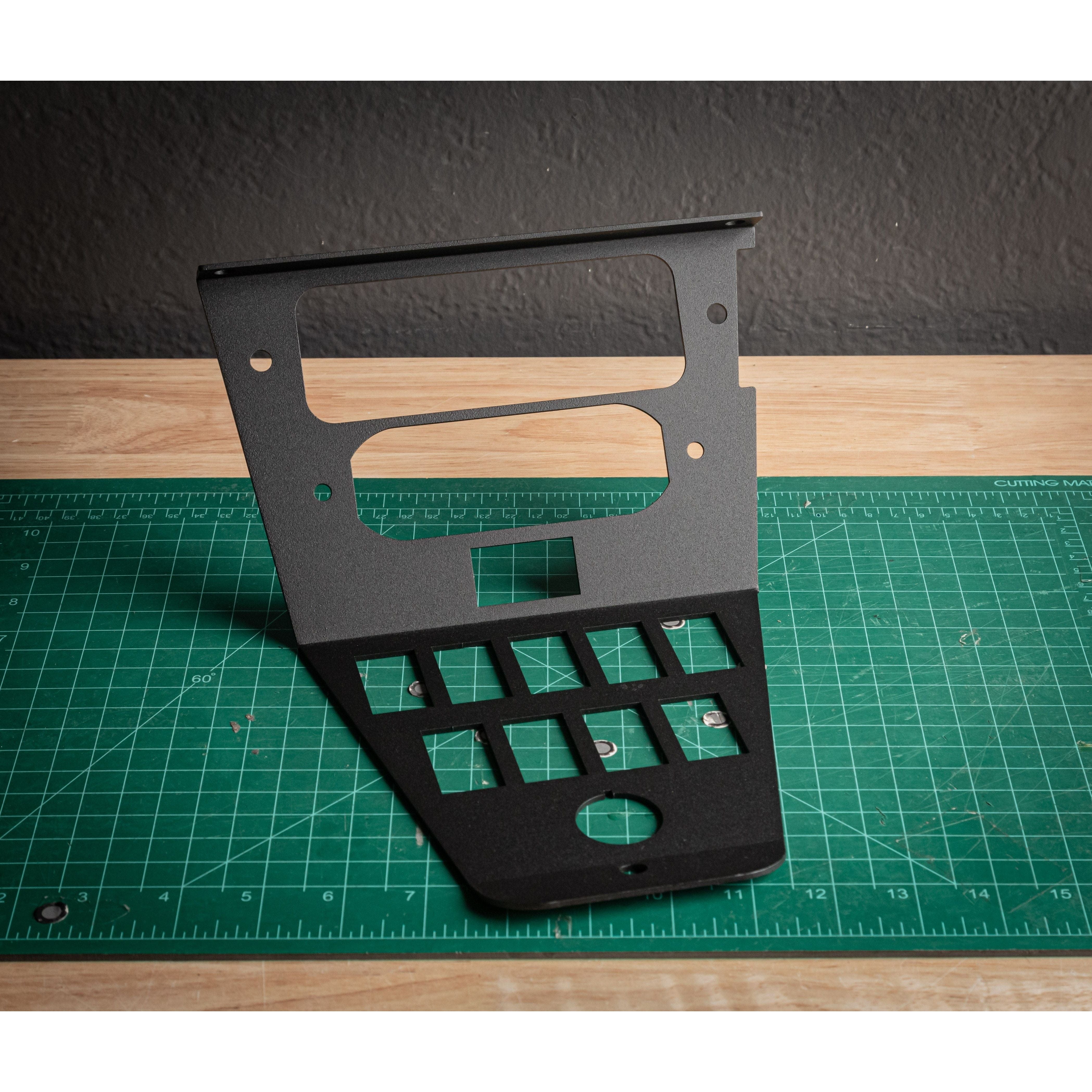 The Polaris ProR/XP RDM Digital Radio Mount FRM by StayFlush, a black metal panel with various cutouts, is shown upright on a green cutting mat with gridlines atop a wooden surface against a dark textured wall.