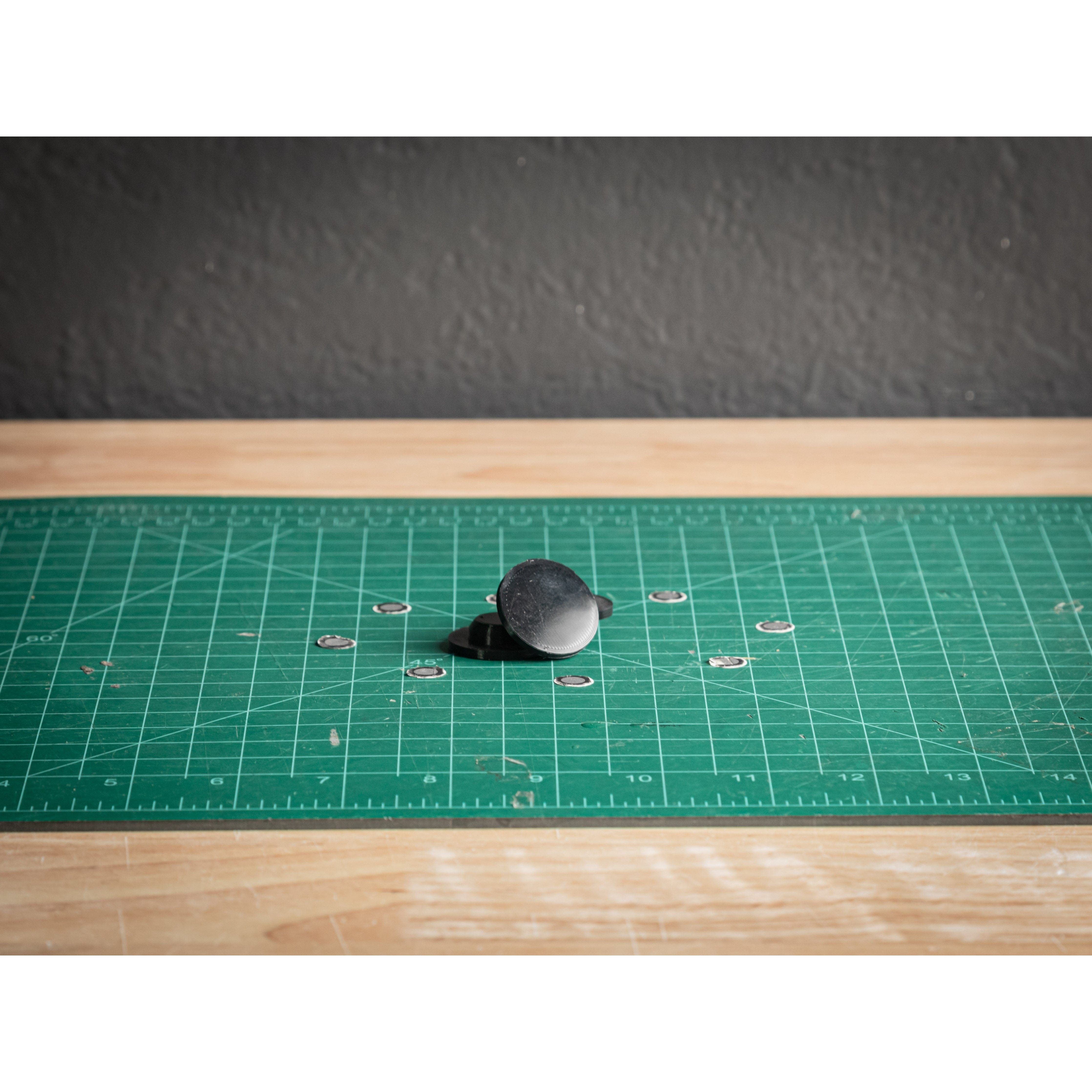 A black and white spinning top sits on a green grid cutting mat, surrounded by small metal washers and PTT Caps by StayFlush, on a wooden table against a dark wall.