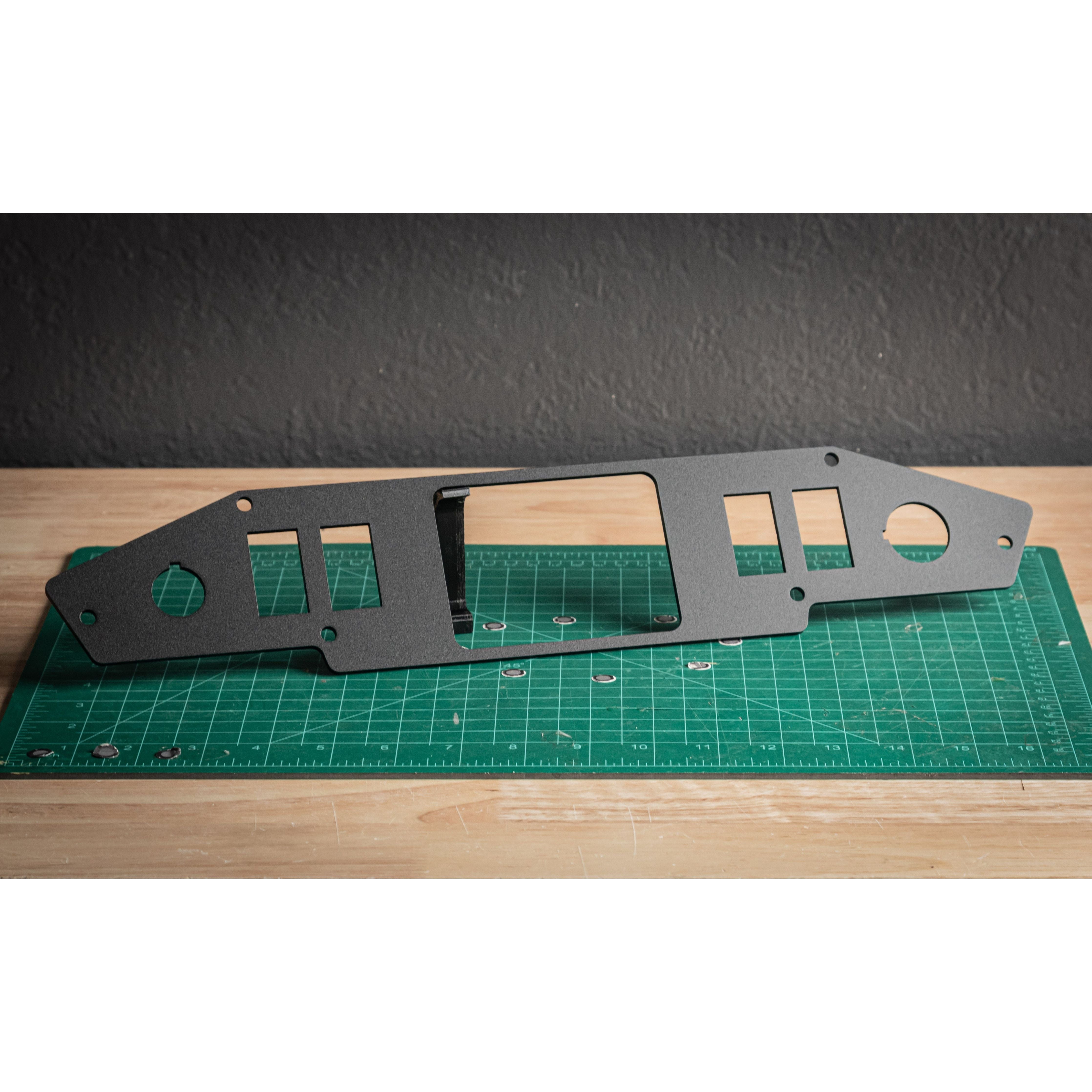 The Turbo S RLB 8 Gang Mount by StayFlush, a black metal dashboard panel with rocker switch slots and cutouts, stands upright on a green cutting mat on a wooden table against a textured gray wall.