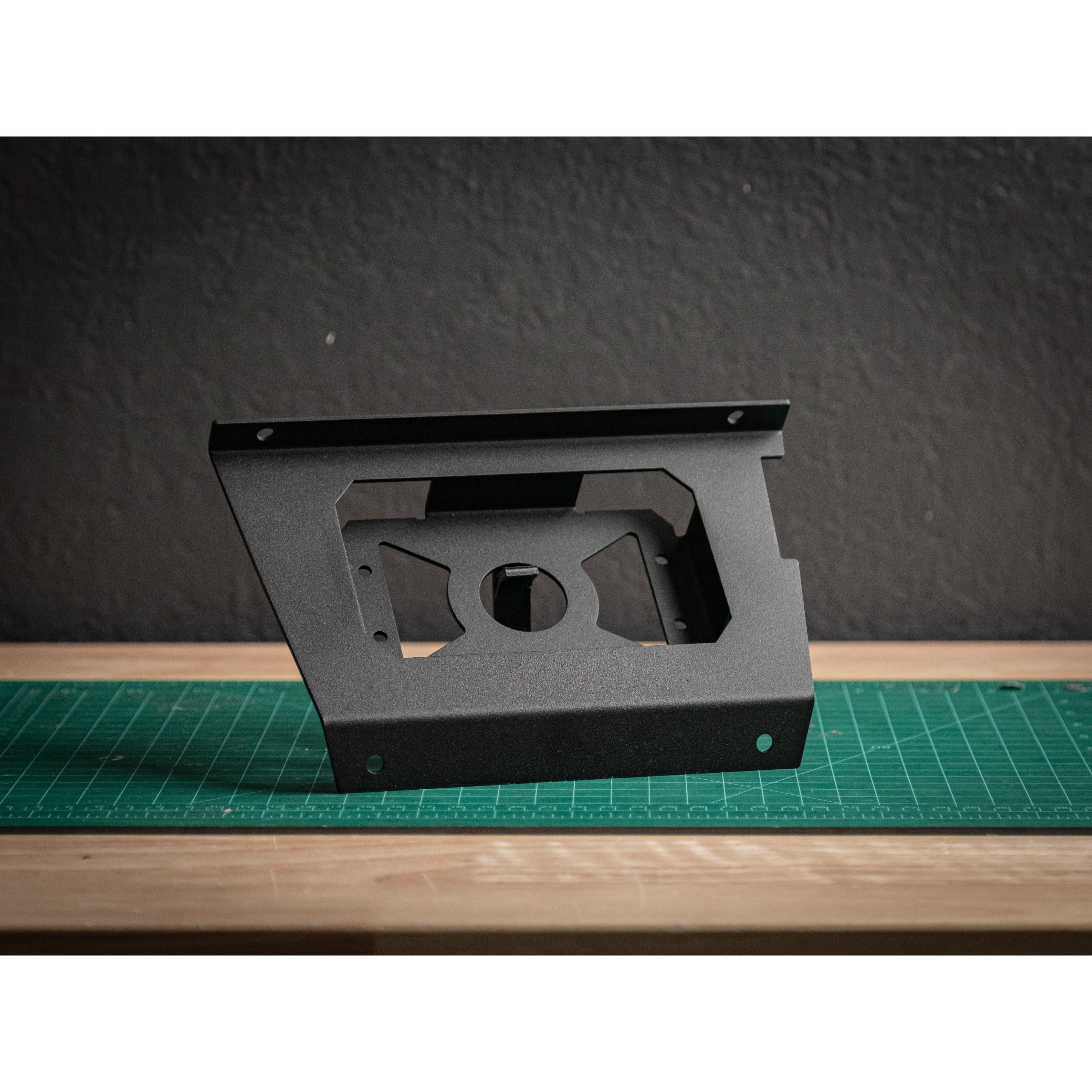 The Pro/R/XP Switchworks Mount by StayFlush, a black metal bracket with a geometric cutout, stands on a green cutting mat atop wood, backed by a dark textured wall for an authentic factory vibe.