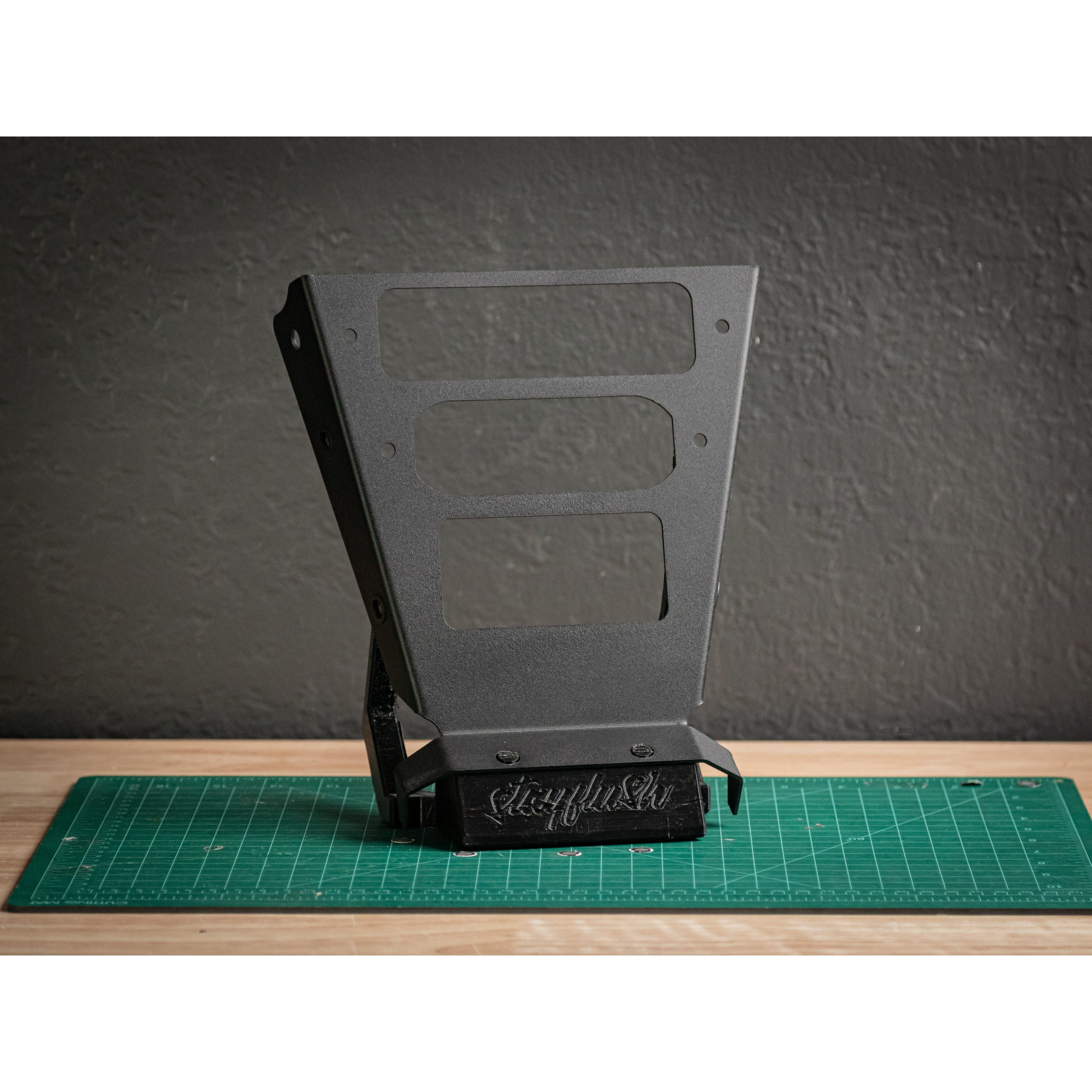 The X3 one off console 0014 by StayFlush is a black metal bracket with rectangular cutouts, perfect for mounting devices such as Rugged Radios or SwitchPro 8, displayed upright on a green cutting mat atop a wooden surface.