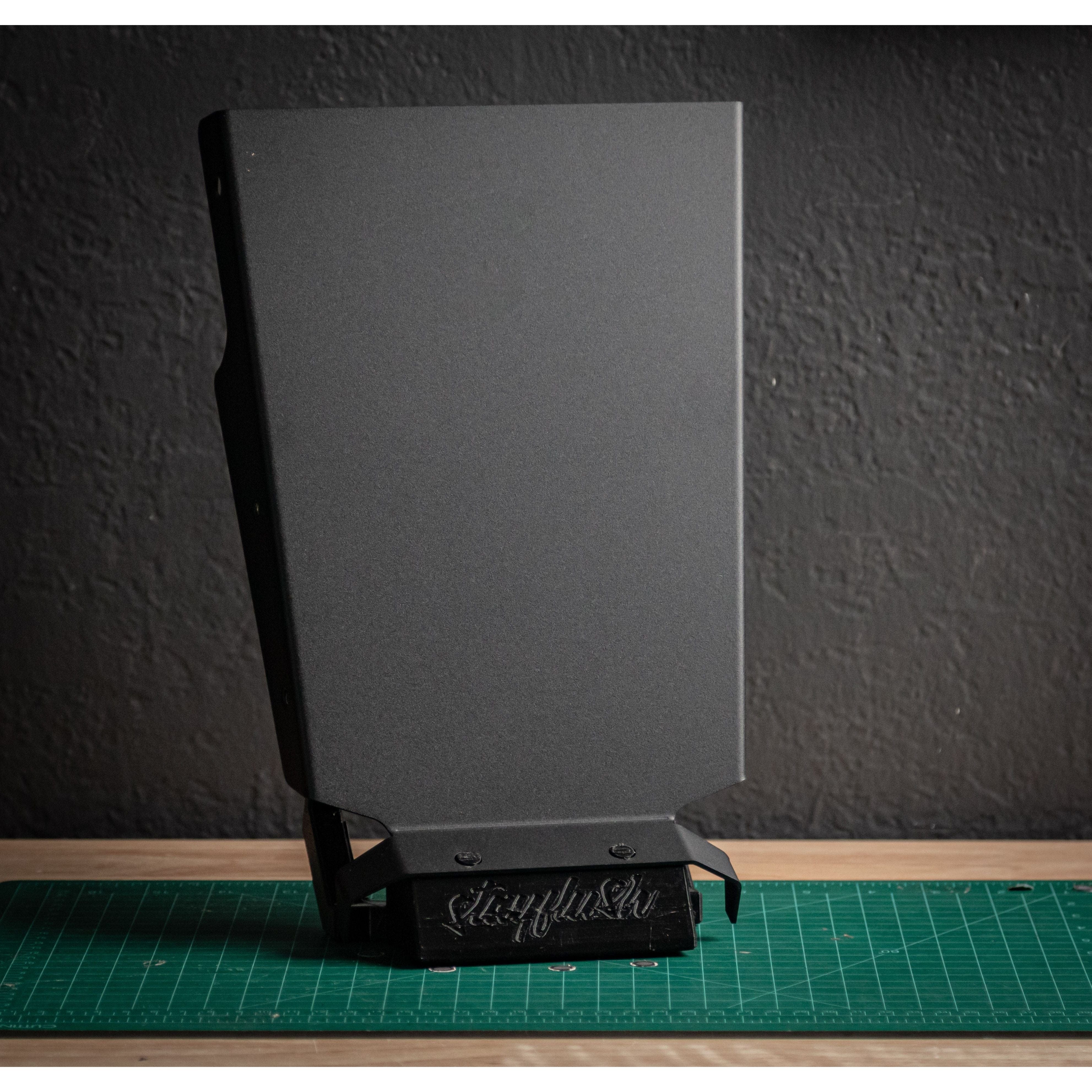 The StayFlush Clean Slate Console XL by StayFlush, a matte black angular metal piece with an engraved logo at the base, stands upright on a green cutting mat. Perfect for customizing console setups or Canamx3 17-25 interiors.
