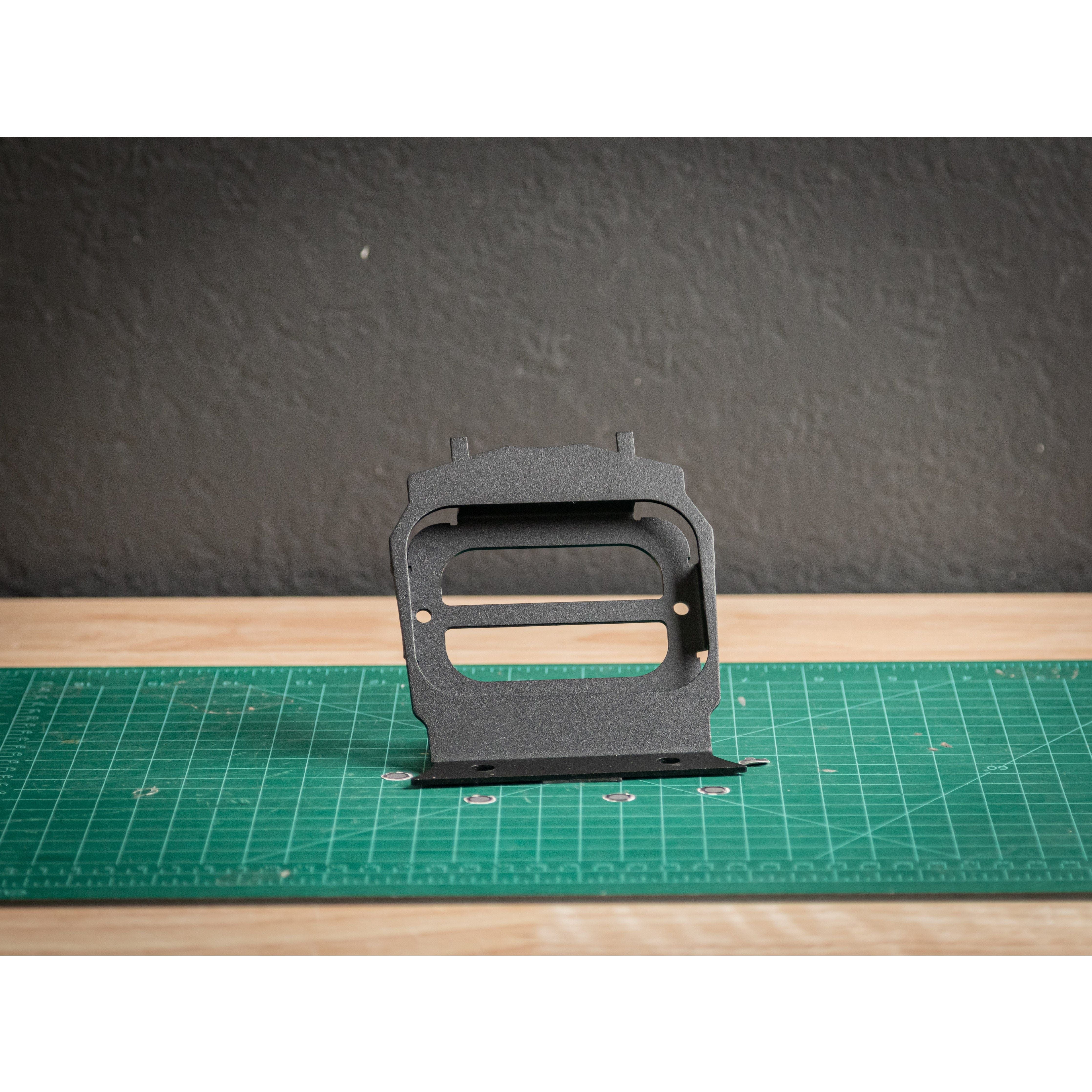 A black StayFlush X3 Lower 6-Gang Auxbeam Mount by StayFlush with rectangular cutouts stands upright on a green cutting mat with grid lines, against a wooden surface and textured dark wall background.